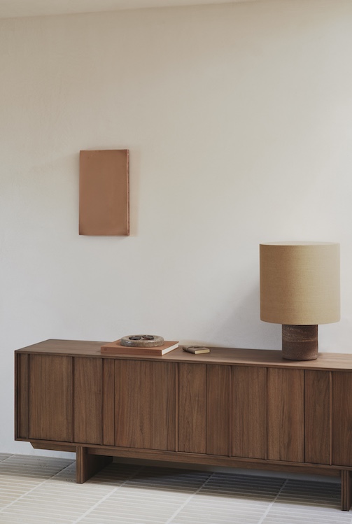 Un buffet en bois de quatuor design avec des livres, un plateau rond et une lampe de table beige est placé contre un mur blanc uni orné d'une petite œuvre d'art rectangulaire. Parfait pour tout amateur de magasin de meubles design.