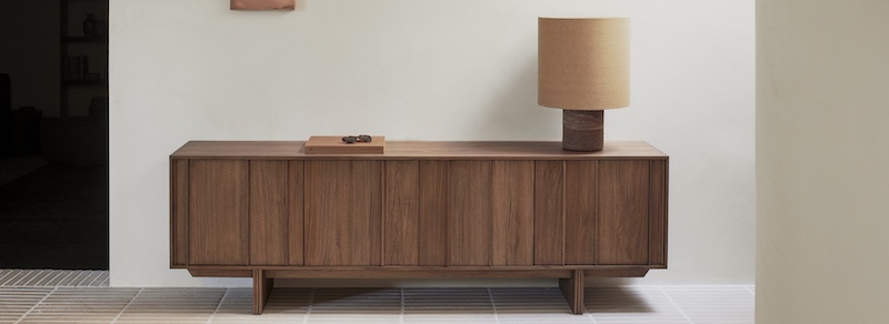 Un buffet en bois, une lampe de table marron clair et un petit plateau sont posés contre un mur blanc uni sur un sol carrelé, apportant une élégance discrète inspirée du design quatuor dans votre maison, parfait pour tout magasin de meubles design.