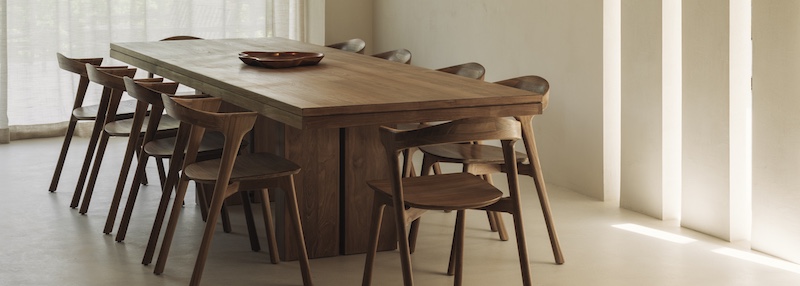 Une longue table de salle à manger en bois de Quatuor Design est entourée de dix chaises assorties dans une pièce minimaliste aux murs neutres et où la lumière du soleil passe à travers des fenêtres verticales, parfaite pour tout magasin de meubles design.