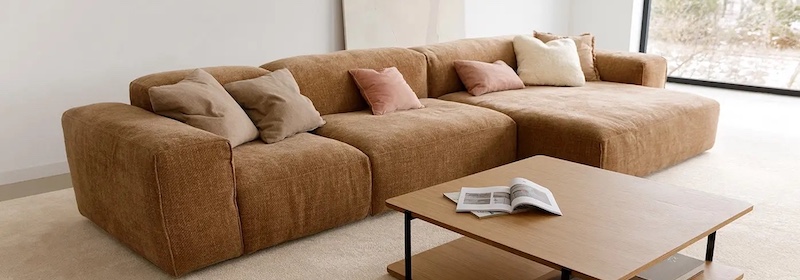 Un grand canapé sectionnel marron d'une collection de design quatuor avec des coussins beiges et roses est disposé dans un salon moderne à côté d'une table basse en bois sur laquelle se trouve un magazine ouvert.