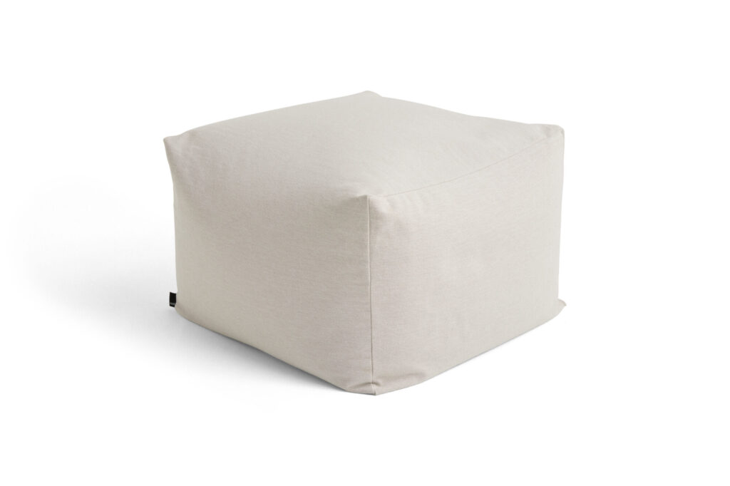 Un pouf ottoman beige clair, recouvert de tissu et aux bords doux, se détache sur un fond blanc. De subtils accents bordeaux ajoutent une touche de chaleur à son design moderne.