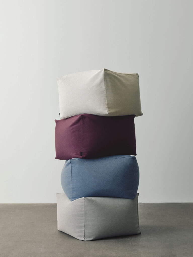 Quatre coussins Pouf en beige, bordeaux, bleu et gris clair sont empilés sur un sol uni contre un mur neutre.