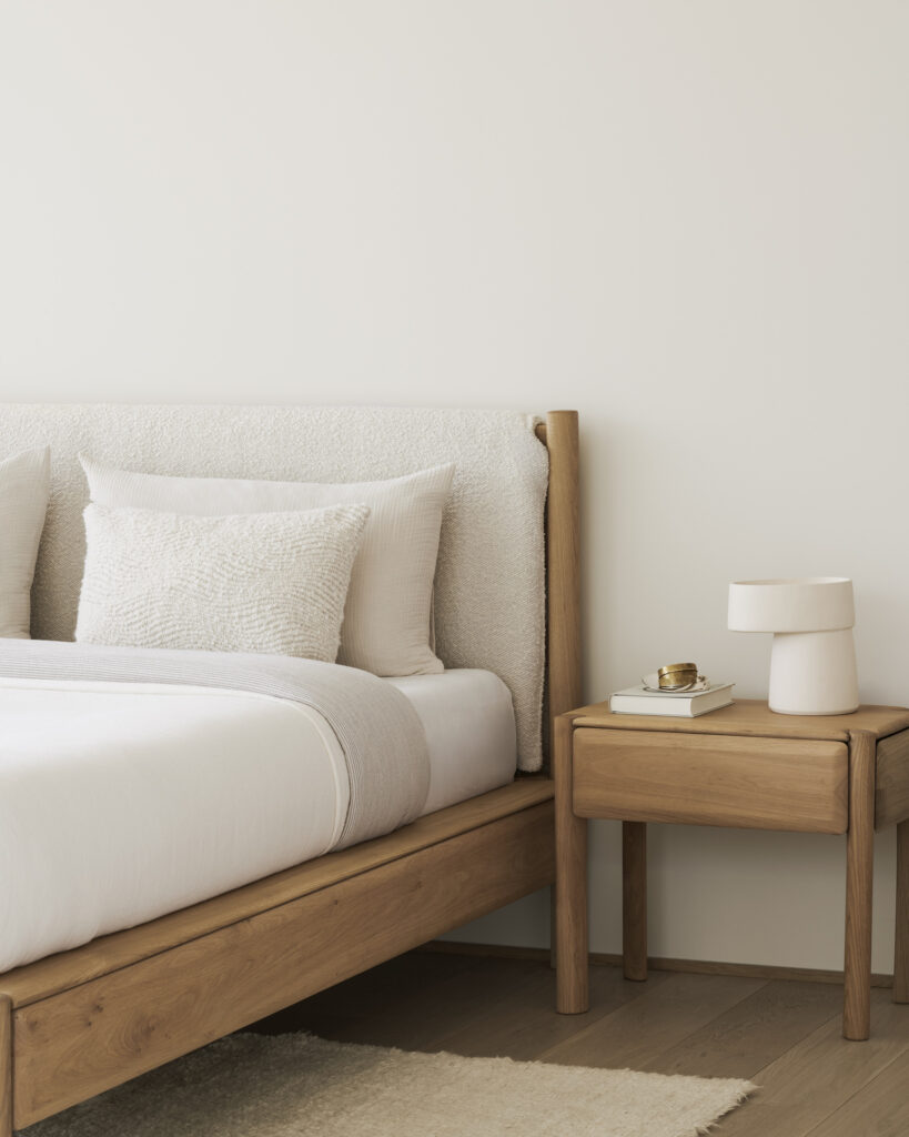 Chambre à coucher minimaliste comprenant un lit PI (160x200) avec literie blanche, une table de nuit en bois contenant une lampe, un livre et un petit plat, le tout posé sur un mur blanc uni pour une ambiance sereine et nuageuse.