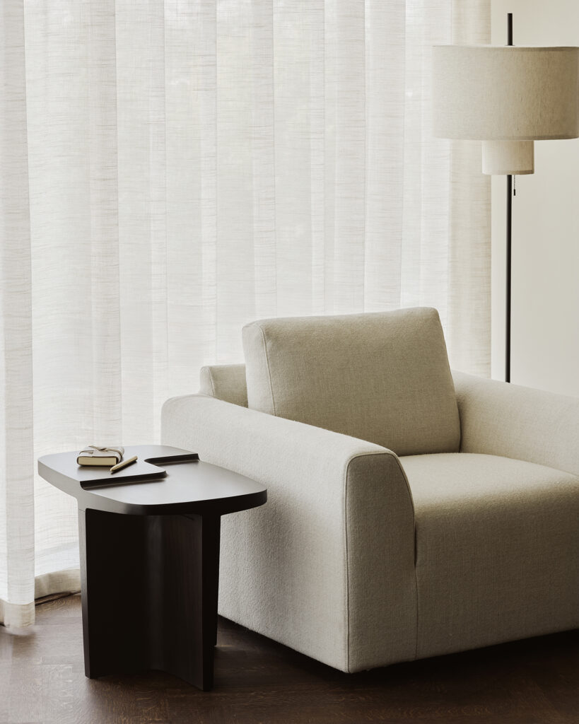 Une chaise longue beige est assise à côté d'une table d'appoint en bois foncé sur laquelle se trouvent un livre fermé et des verres ; un lampadaire et des rideaux transparents se trouvent à l'arrière-plan.