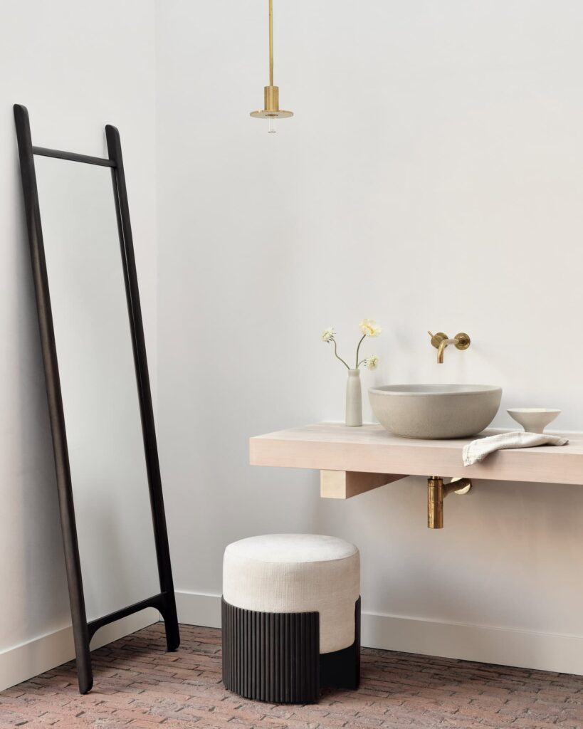 Salle de bains minimaliste avec un lavabo rond sur un meuble-lavabo flottant en bois, un miroir PI au sol, un petit tabouret, une échelle noire, une lampe suspendue et deux petits vases avec des fleurs sur des murs blancs.