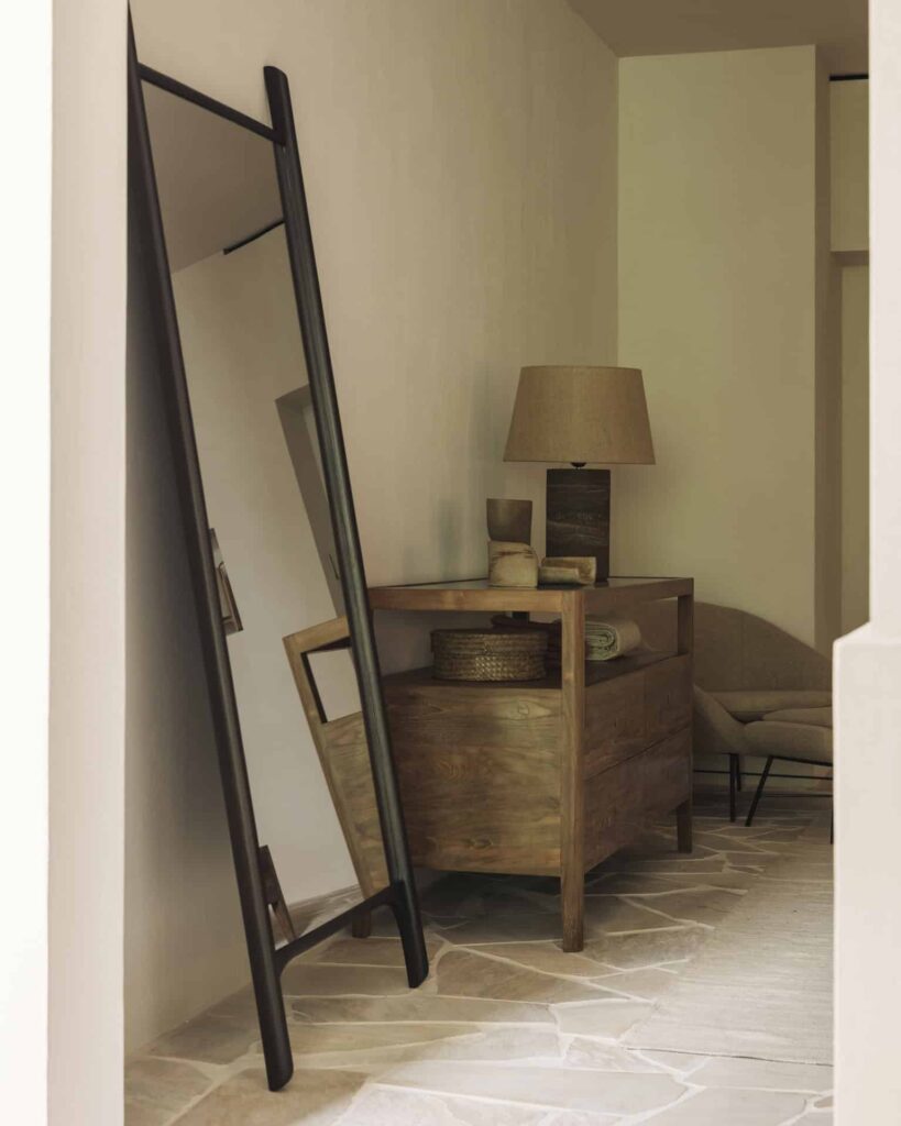 Un grand miroir de sol PI avec un cadre brun foncé est adossé à un mur à côté d'un buffet en bois surmonté d'une lampe, de paniers et de céramiques dans une pièce à la décoration minimaliste.
