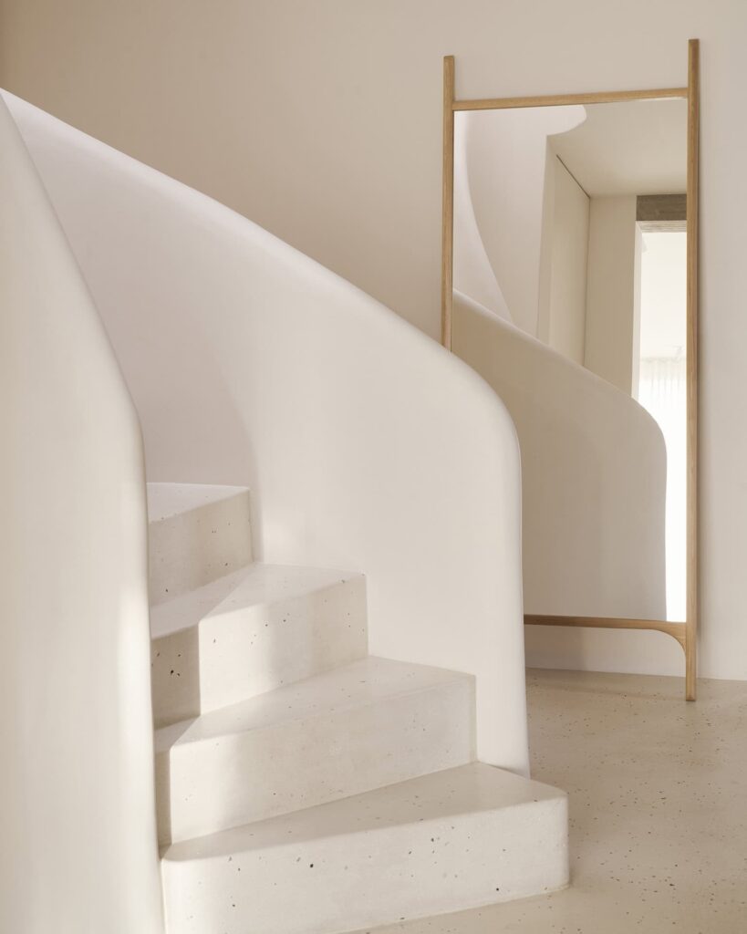 Un escalier en colimaçon blanc minimaliste se reflète dans le grand miroir de sol PI avec un cadre en chêne naturel, placé sur un mur et un sol aux tons neutres.