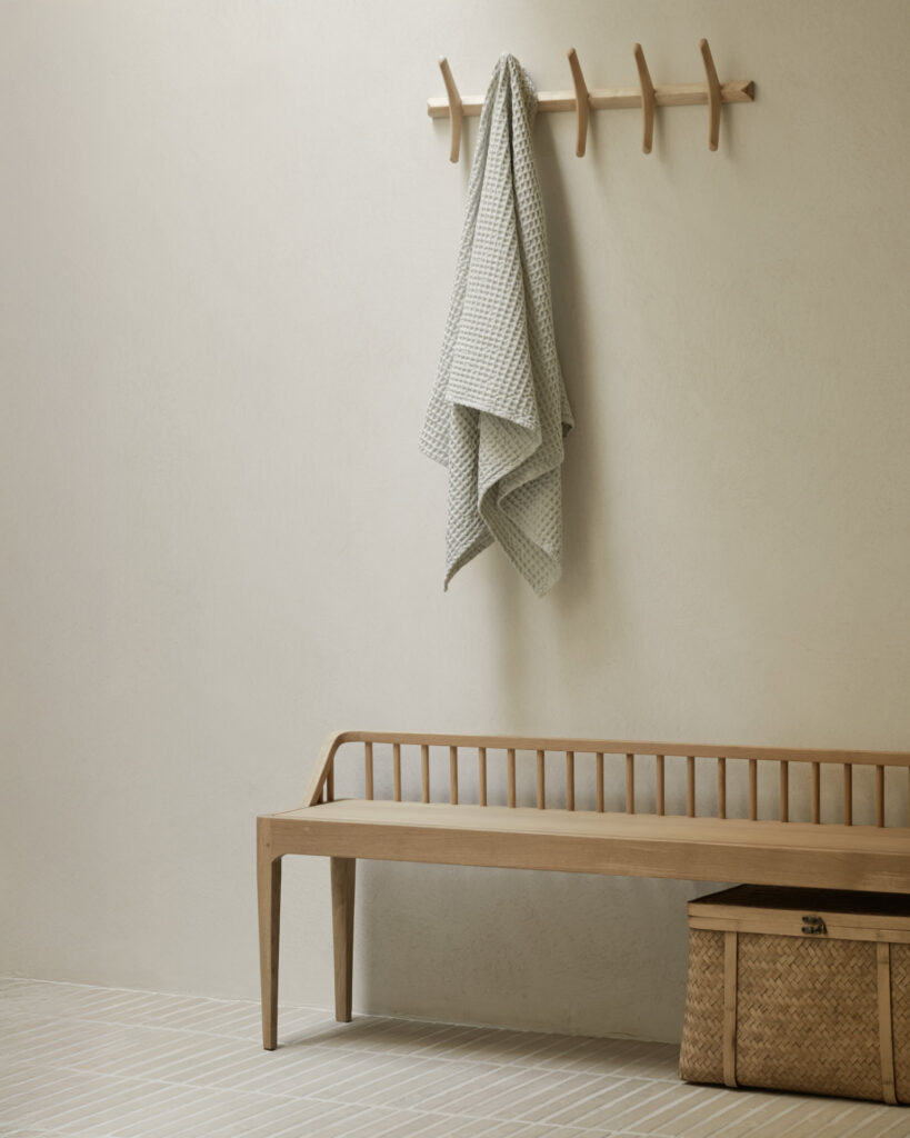 Un banc en bois clair est posé contre un mur beige avec une serviette suspendue à une patère murale au-dessus ; un panier tressé est posé à côté du banc sur un sol rayé.
