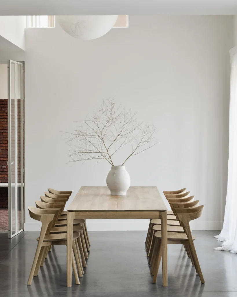 Une salle à manger minimaliste avec une table à rallonge en chêne naturel verni, huit chaises assorties et un vase blanc contenant des branches nues, sur un mur blanc éclairé par la lumière naturelle.