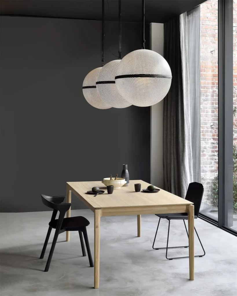 Une salle à manger minimaliste avec une table en bois clair chêne naturel à rallonge, trois chaises noires, et trois grandes lampes suspendues sphériques, placées contre des murs sombres et une grande fenêtre avec une vue sur l'extérieur en briques.