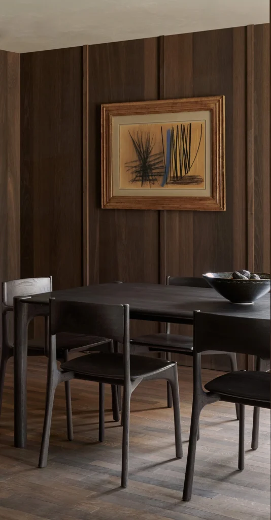 Une salle à manger minimaliste avec des murs en bois brun foncé verni, une table noire avec quatre chaises PI, un bol sur la table et une œuvre d'art abstraite encadrée au mur.