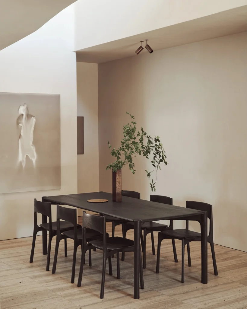 Salle à manger minimaliste avec une table brun foncé, huit chaises PI, un vase avec des branches vertes, un sol en bois clair et un art mural texturé.