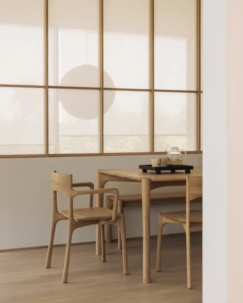Un coin repas minimaliste avec des chaises PI en bois clair et en chêne naturel, un plateau noir avec des tasses et une théière, et de grandes fenêtres avec des rideaux transparents en arrière-plan.