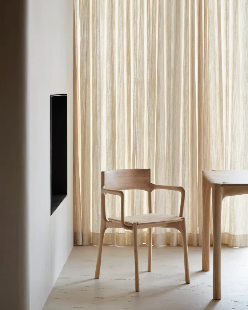 Une chaise PI en bois avec accoudoirs et une table en chêne naturel trônent dans une pièce minimaliste avec des rideaux clairs et une niche murale encastrée.