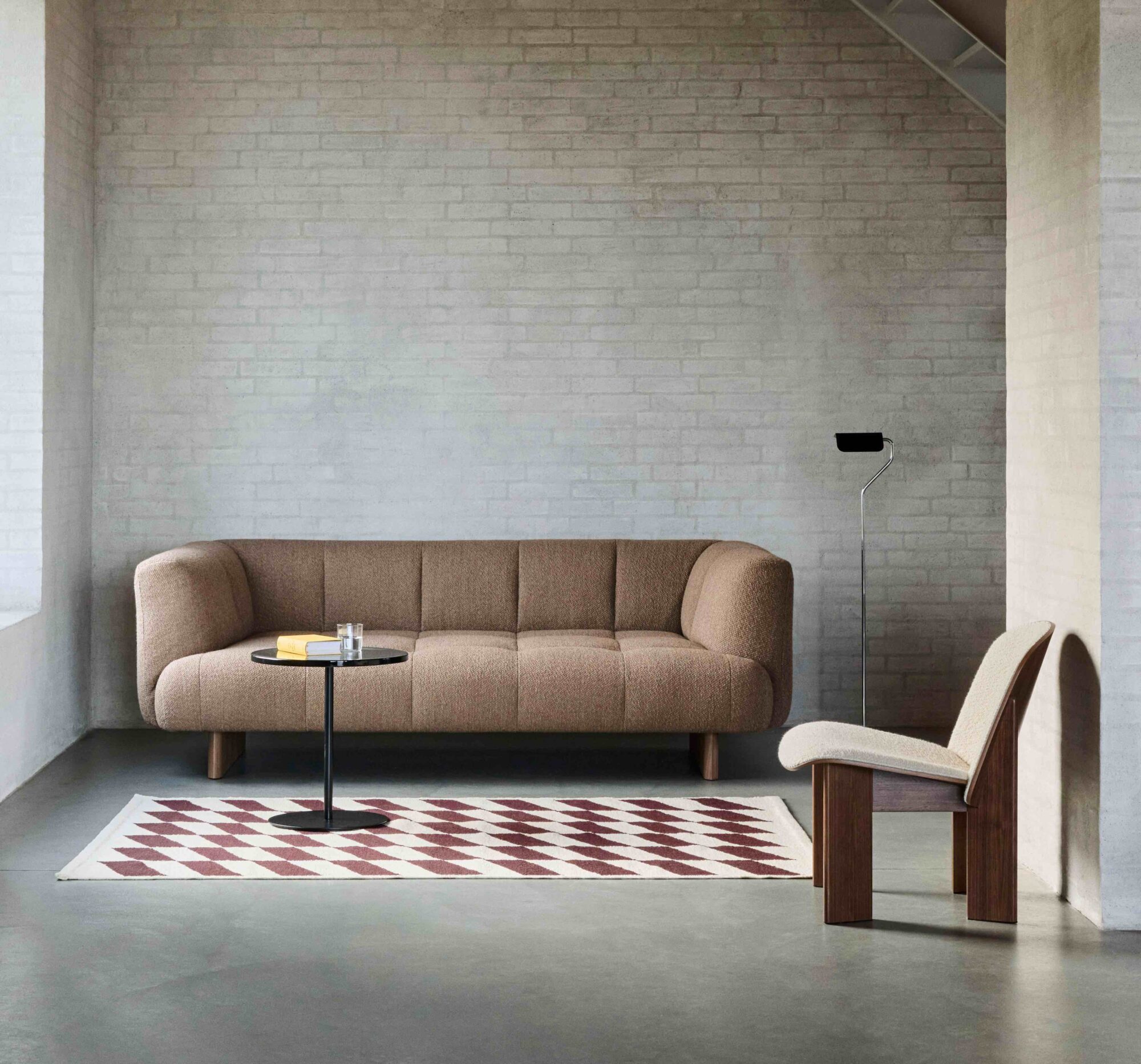 Un salon minimaliste avec un canapé marron, une chaise en bois et une table basse ronde noire, complété par un tapis à motifs géométriques sur un sol en béton - une inspiration parfaite du design quatuor ou de votre magasin de meubles design local.