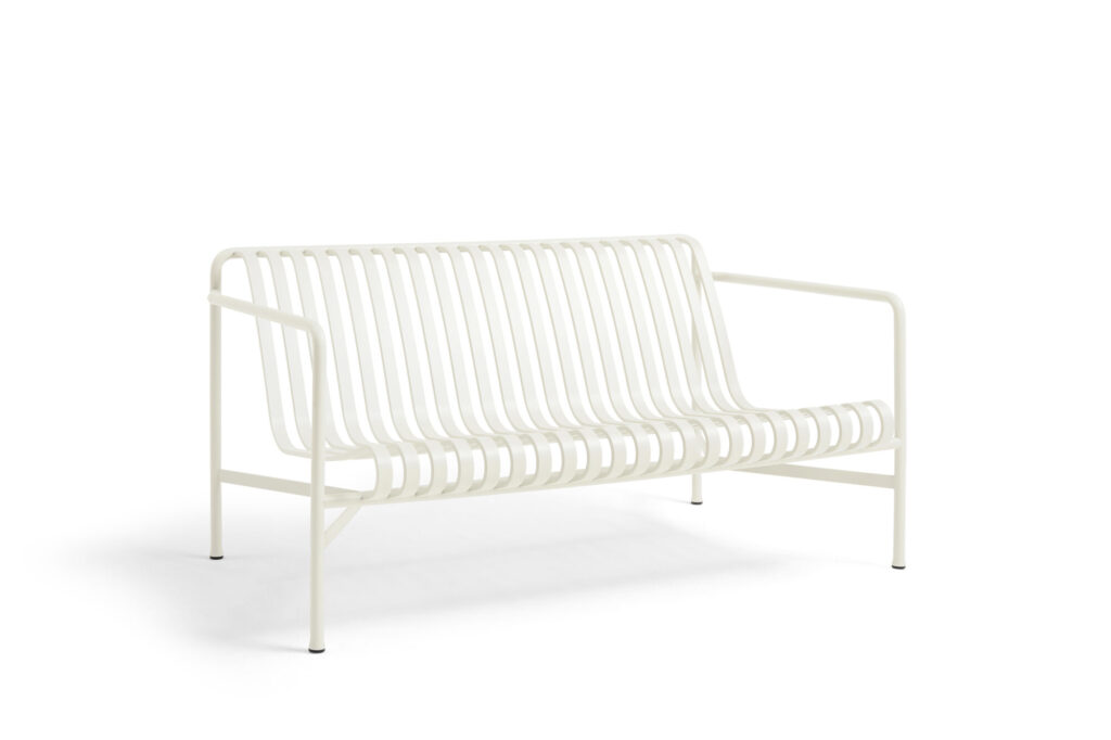 Une banquette en métal blanc crème aux lattes régulièrement espacées, dotée d'accoudoirs et de quatre pieds droits, évoque le style minimaliste d'un Sofa Palissade, sur un fond blanc uni.