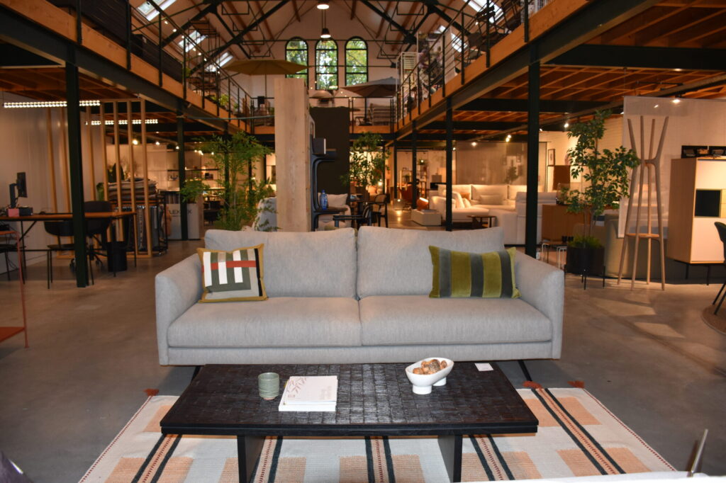 Un showroom moderne de style loft présente un Canapé Diva 3 places, un canapé tissu Colourwash avec coussins, une table basse noire et un décor de style industriel avec des poutres apparentes et des plantes.