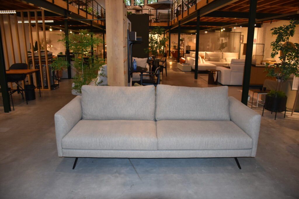 Un Canapé Diva 3 places gris clair, tissu Colourwash avec de fins pieds en métal se trouve au centre d'une salle d'exposition de meubles spacieuse et moderne.