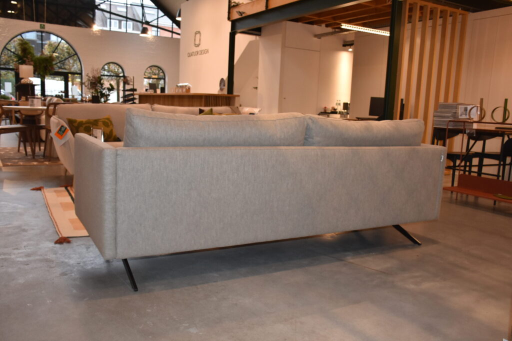 Un Canapé Diva 3 places, tissu Colourwash, avec des pieds en métal est placé au centre d'une salle d'exposition de meubles avec de grandes fenêtres et un décor moderne.