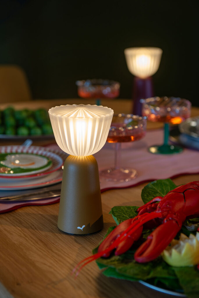 Une Lampe Swiing dorée avec un abat-jour plissé blanc brille sur une table à manger composée d'assiettes, de boissons, de pains d'épices et d'un homard cuit.