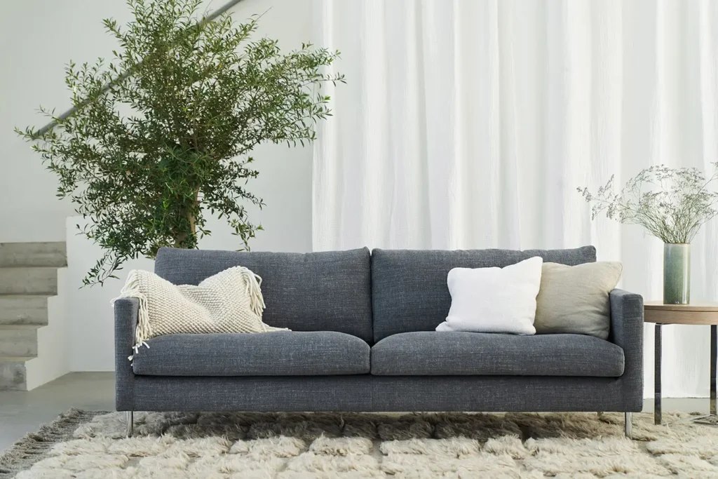 Un salon moderne avec un canapé gris foncé en stock, trois coussins, une couverture beige, une plante en pot, une petite table avec un vase et un tapis texturé au sol.