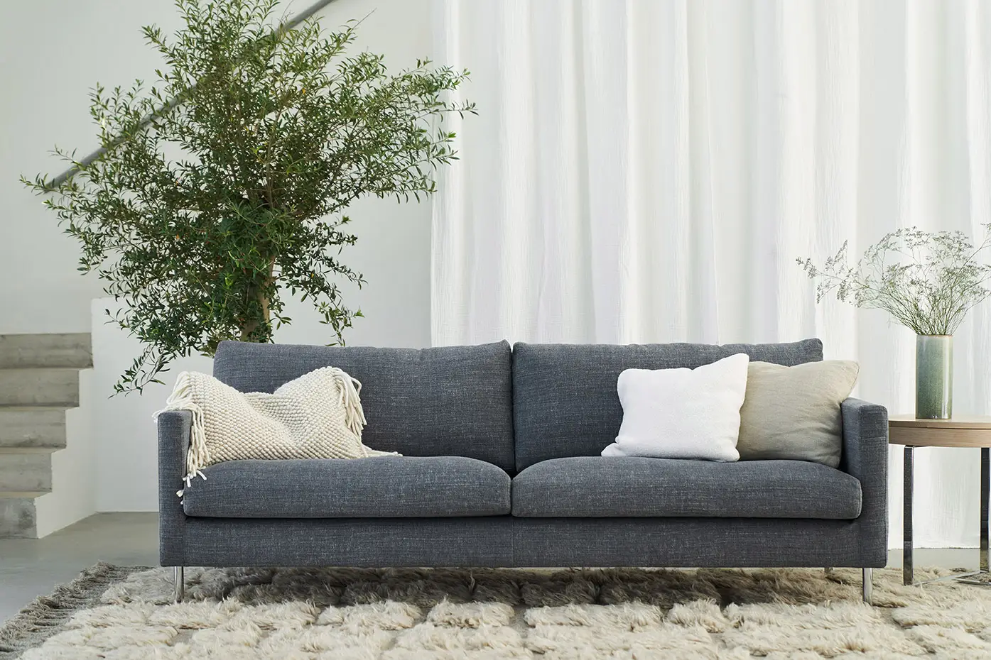Un salon moderne avec un canapé gris foncé en stock, trois coussins, une couverture beige, une plante en pot, une petite table avec un vase et un tapis texturé au sol.