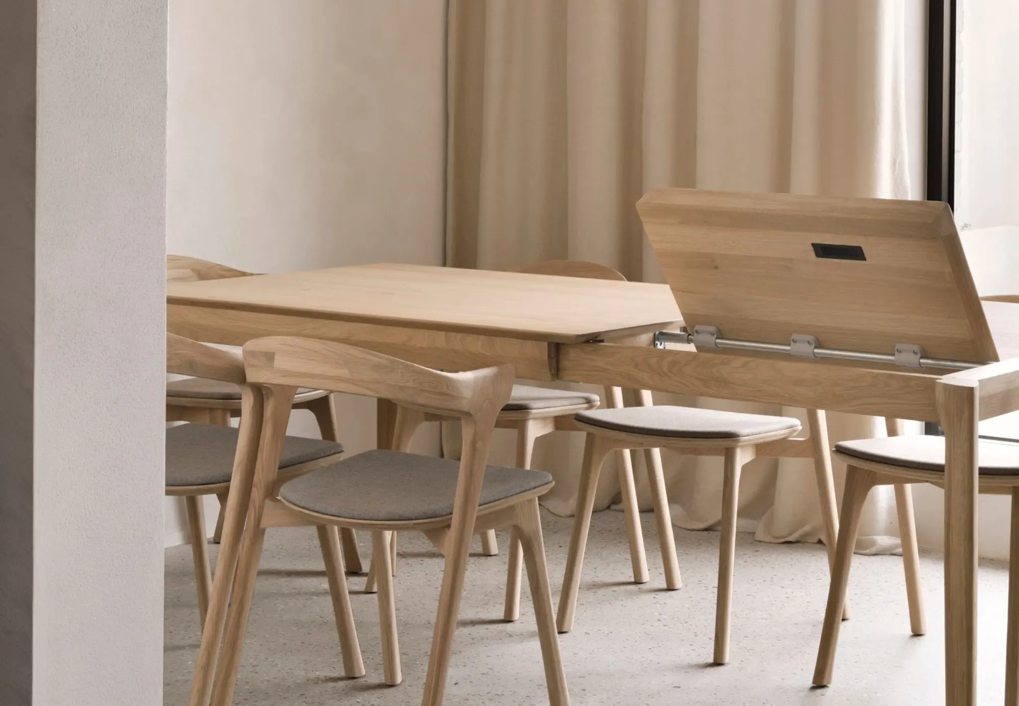 Une table de salle à manger en bois clair dont l'un des côtés se soulève pour révéler un espace de rangement, parfaite pour optimiser vos espaces, entourée de chaises assorties avec des coussins gris clair, le tout dans une pièce minimale aux tons neutres.