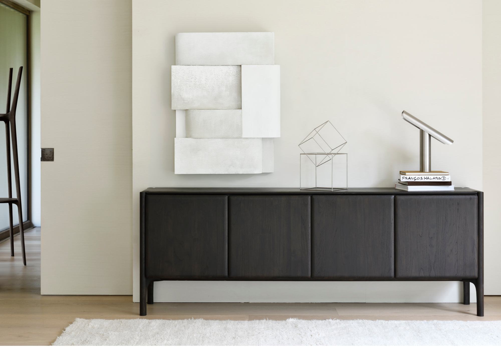 Un buffet moderne en bois foncé se dresse contre un mur clair, surmonté de sculptures abstraites, d'un livre et d'une lampe de bureau argentée - parfait pour optimiser vos espaces pour les fêtes - avec des œuvres d'art géométriques au-dessus.