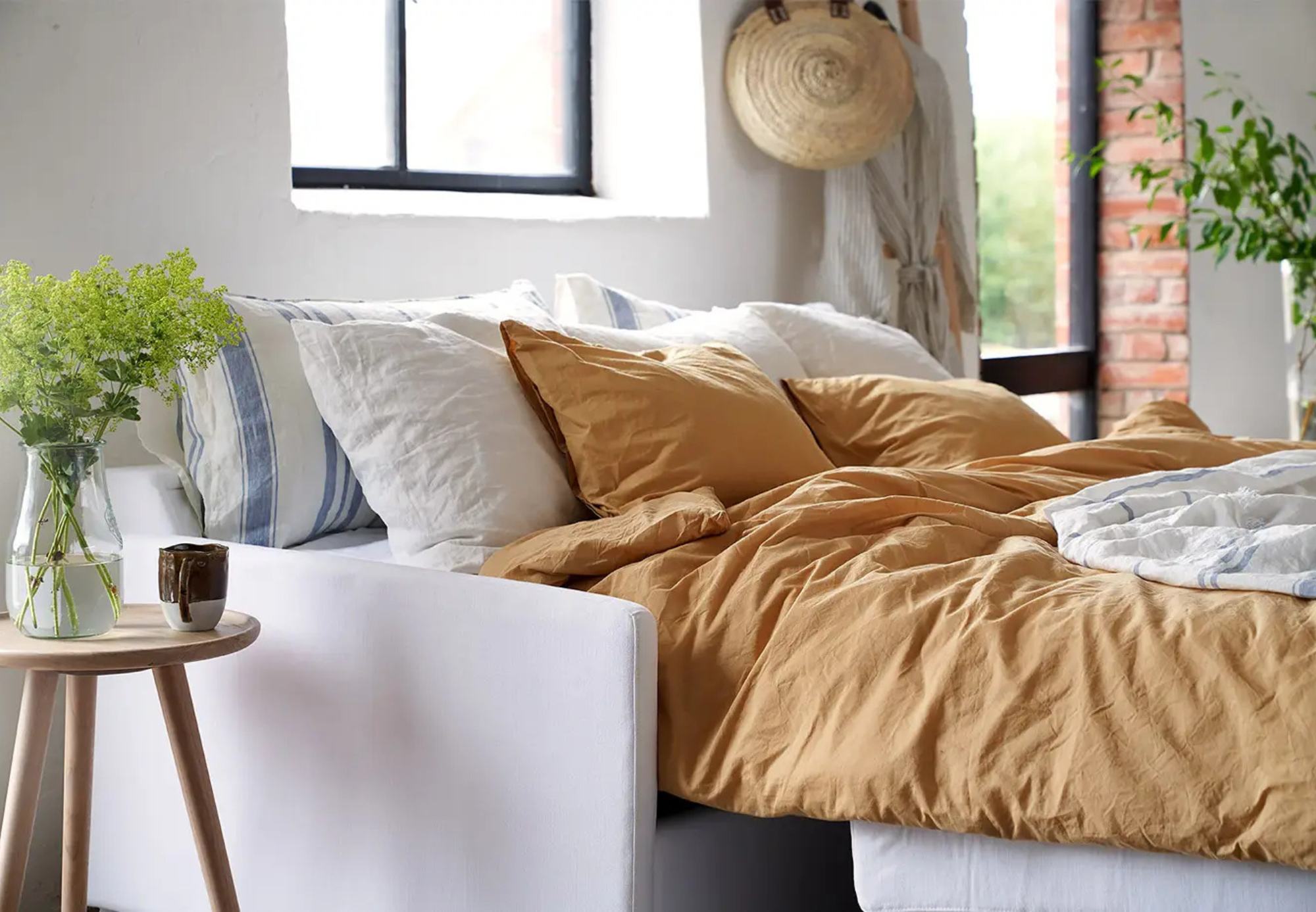 Une chambre à coucher moderne avec un cadre de lit blanc, une literie jaune moutarde, des oreillers rayés et une table d'appoint avec un vase de fleurs vertes est conçue pour optimiser les espaces, remplis de lumière naturelle provenant d'une grande fenêtre.