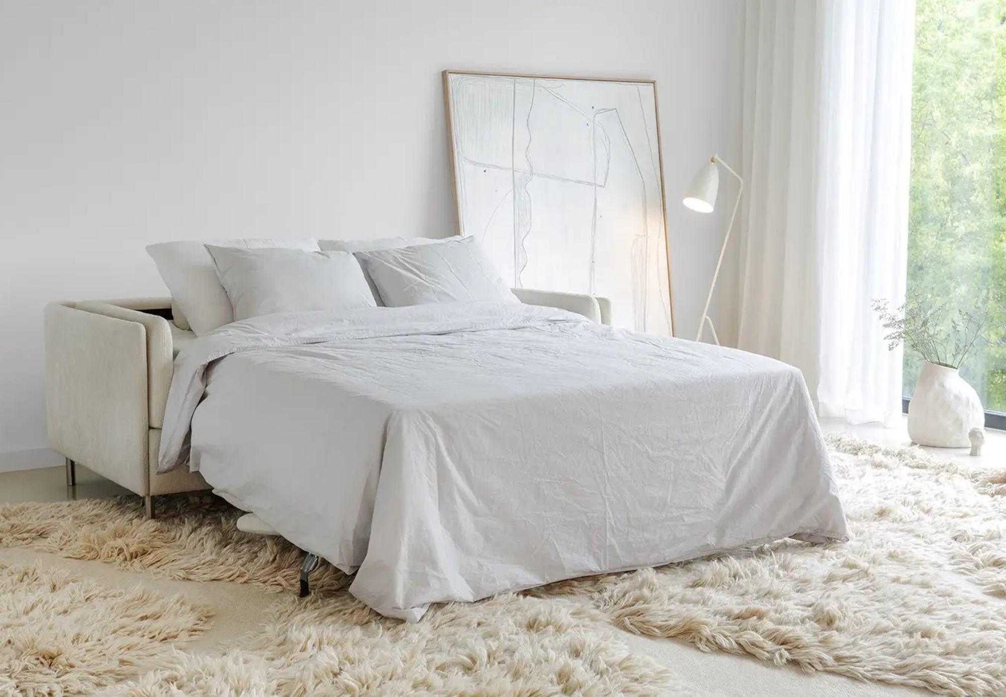 Un lit soigneusement fait avec des draps blancs est posé sur un tapis à poils longs dans une chambre lumineuse, où de grandes fenêtres et des œuvres d'art abstraites contribuent à optimiser les espaces, complétés par un lampadaire et un vase en céramique.