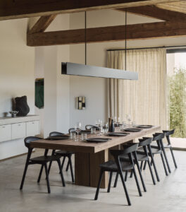 Une salle à manger moderne avec une longue table en bois pour huit personnes, des chaises noires, une décoration minimaliste et des poutres apparentes - parfaite pour optimiser les espaces et accueillir des fêtes - baignée par la lumière naturelle d'une fenêtre aux rideaux beiges.