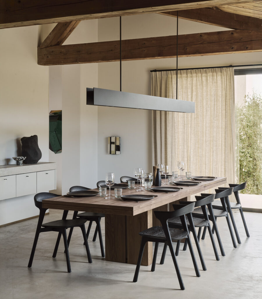 Une salle à manger moderne avec une longue table en bois pour huit personnes, des chaises noires, une décoration minimaliste et des poutres apparentes - parfaite pour optimiser les espaces et accueillir des fêtes - baignée par la lumière naturelle d'une fenêtre aux rideaux beiges.